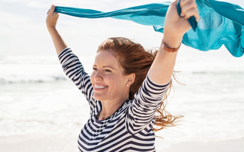 red hair woman smiling at beach holding scarf up in wind